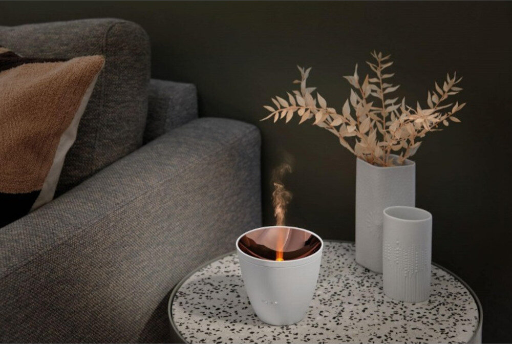 Na okrągłym stoliku z wzorem w kropki stoi biały aromatyzer z widoczną parą. Obok znajdują się dwa wazoniki z suchymi gałązkami.