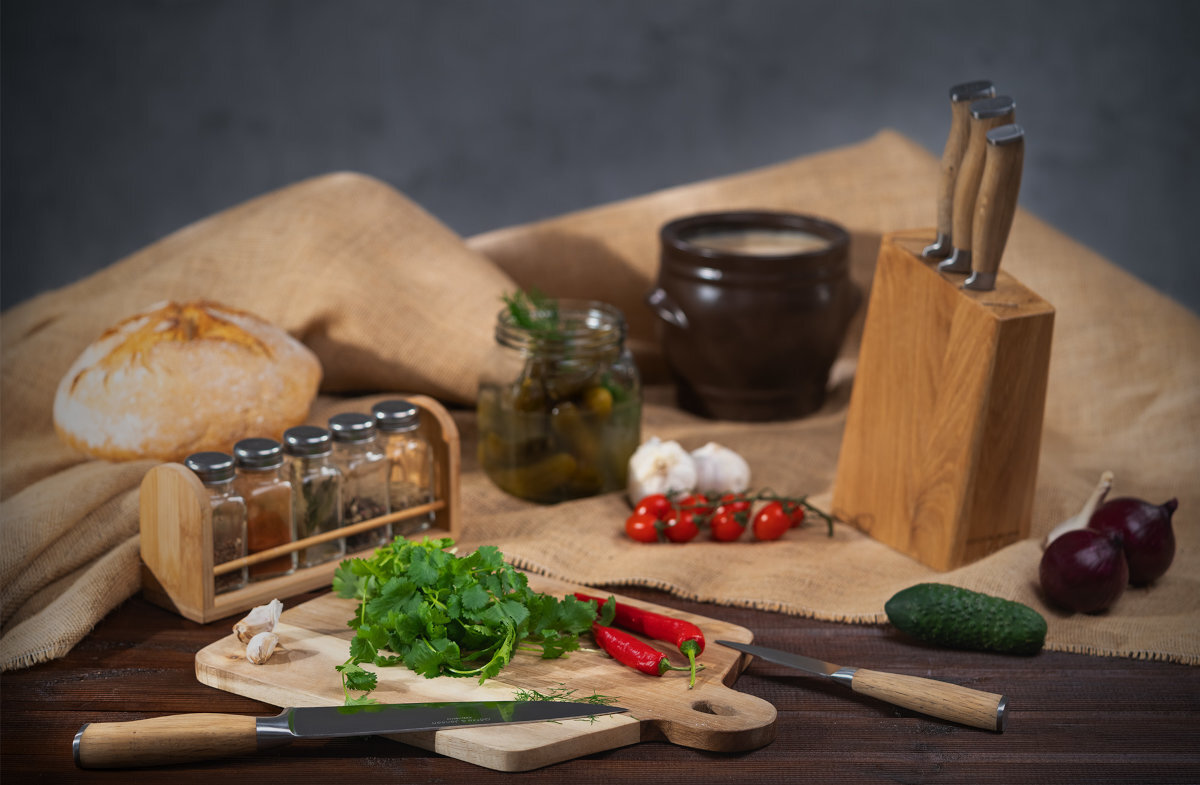 Na drewnianym blacie znajduje się zestaw noży w bloku z drewna, deska do krojenia z ziołami i papryczkami oraz stojak z przyprawami. W tle widoczne są chleb, słoik z ogórkami, czosnek, pomidorki koktajlowe, cebula i gliniany garnek.