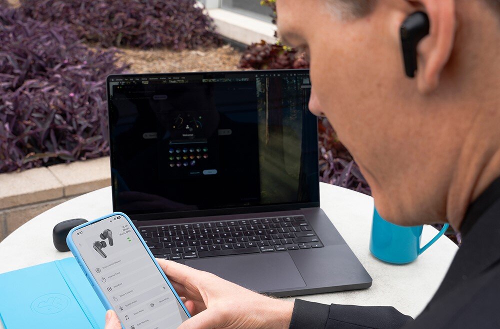Słuchawki dokanałowe JLAB Jbuds Pods ANC, dedykowana aplikacja, personalizacja dźwięku, osoba trzymająca smartfon w rękach siedząca przy stoliku z laptopem