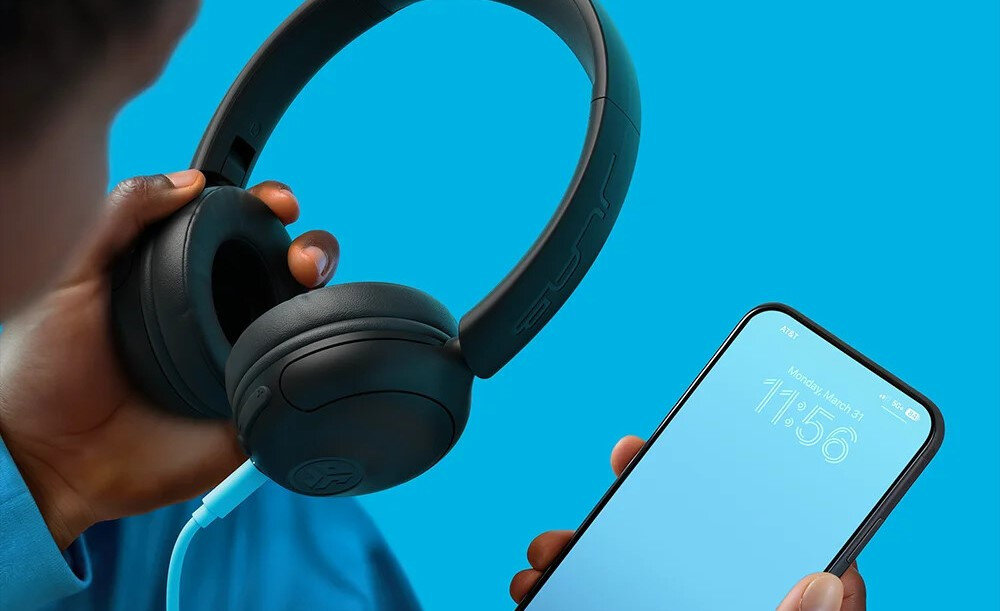 Słuchawki nauszne JLAB Headest STUDIO 2 Bluetooth bezprzewodowe połączenie muzyka rozmowy, osoba trzymająca w ręku słuchawki oraz smartfon