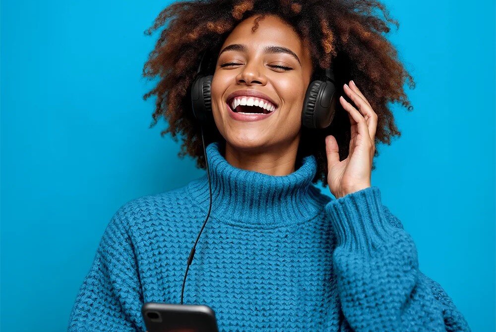 Słuchawki nauszne JLAB Wired Headset STUDIO 2, wyraźny dźwięk stereo, komfort pracy, osoba z założonymi sluchawkami i smartfonem w ręku, która się uśmiecha