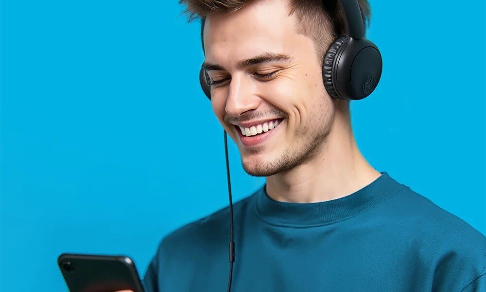 Słuchawki nauszne JLAB Wired Headset STUDIO 2, dynamiczne membrany, pasmo 20-20000 Hz, osoba z założonymi sluchawkami i smartfonem w ręku, która się uśmiecha