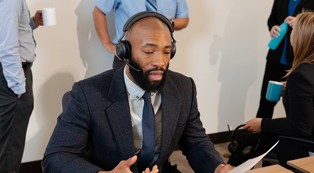 Słuchawki nauszne JLAB Epic Work, lekka konstrukcja, komfort noszenia, osoba z założonymi słuchawkami nausznymi trzymająca kartkę w ręku, osoby stojące z kubkami na drugim planie