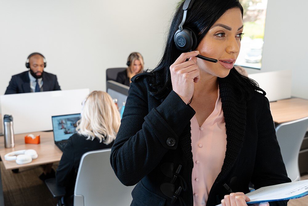 Słuchawki nauszne JLAB Epic Work, akumulator 600 mAh, czas pracy do 90h, osoba stojąca przy biurku z założonymi słuchawkami nausznymi