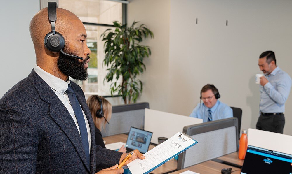 Słuchawki nauszne JLAB Epic Work, kompatybilne z PC i telefonami, osoba stojąca przy biurku z założonymi słuchawkami nausznymi prowadząca rozmowę