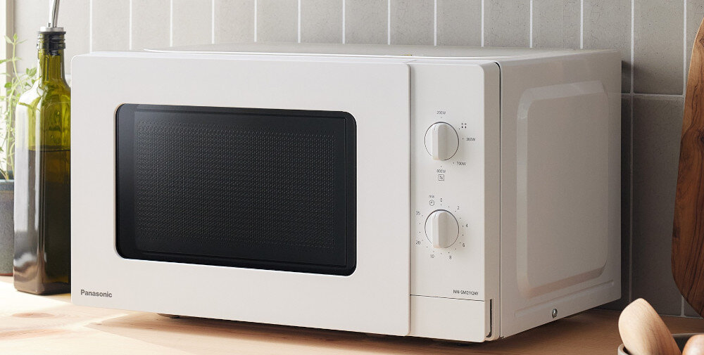KUCHNIA MIKROFALOWA PANASONIC NN-SM21QWEPG Biała kuchenka mikrofalowa Panasonic ze sterowaniem pokrętłami stoi na jasnym drewnianym blacie, otoczona z lewej strony ziołami i oliwą, a z prawej deską z pieczywem i pojemnikiem na przybory kuchenne, tworząc spójny i przytulny wystrój kuchni szybkie podgrzewanie łatwe posiłki ekspresowe rozmrażanie kompaktowy design minimalistyczne pokrętła mała kuchnia seria SM prosta obsługa codzienne użytkowanie