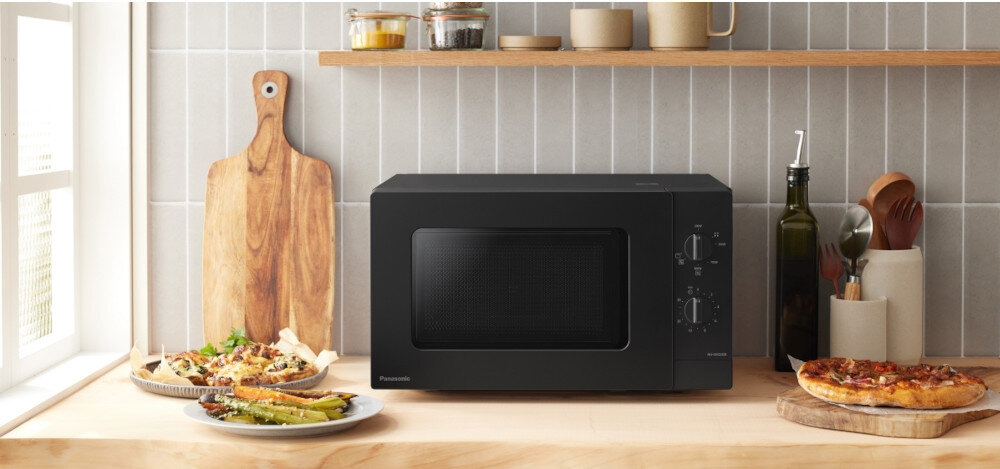 KUCHNIA MIKROFALOWA PANASONIC NN-GM22QBEPG Czarna kuchenka mikrofalowa Panasonic ustawiona na blacie w przytulnej kuchni, otoczona deską do krojenia, oliwą, naczyniami i gotowymi daniami, podkreśla jej elegancki wygląd i dopasowanie do wnętrza.