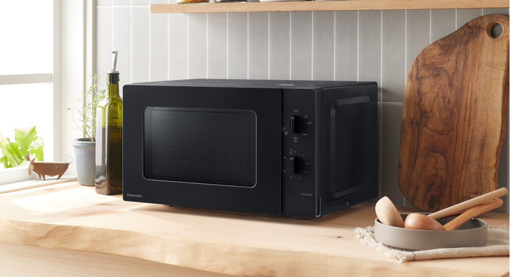 KUCHNIA MIKROFALOWA PANASONIC NN-GM22QBEPG Czarna kuchenka mikrofalowa Panasonic ustawiona na drewnianym blacie przy oknie, w otoczeniu oliwy, rośliny i drewnianej deski, prezentuje swój matowy front do eleganckiej kuchni funkcja grilla chrupiące potrawy szybkie gotowanie seria GT kompaktowy design wygląd mała kuchnia łatwa obsługa codzienny komfort grillowanie w domu