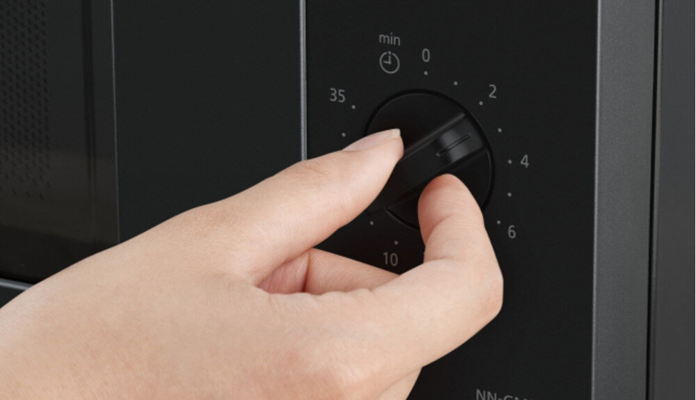 KUCHNIA MIKROFALOWA PANASONIC NN-GM22QBEPG Zbliżenie dłoni przekręcającej pokrętło minutnika w czarnej kuchence mikrofalowej, podkreślające prostą, intuicyjną regulację czasu pracy urządzenia prosta obsługa szybkie gotowanie regulacja mocy ustawienie czasu minimalistyczny panel czytelne pokrętła intuicyjna obsługa wygodne sterowanie codzienny komfort