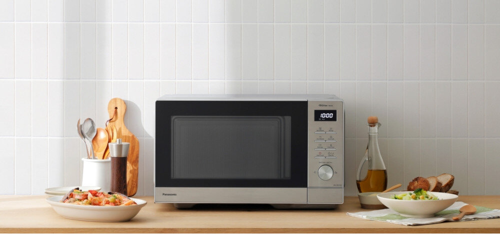 KUCHNIA MIKROFALOWA PANASONIC NN-SD37QSEPG Kuchenka mikrofalowa Panasonic na kuchennym blacie, ustawiona centralnie, otoczona gotowymi daniami, deskami i przyprawami, na tle białych płytek ściennych, prezentująca elegancki design urządzenia w aranżacji kuchni