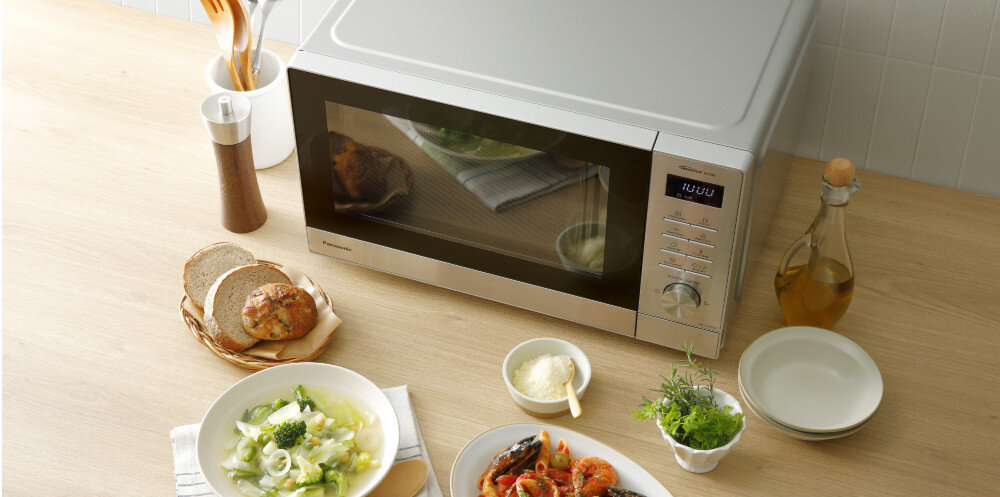 KUCHNIA MIKROFALOWA PANASONIC NN-SD37QSEPG Widok z góry na kuchenkę mikrofalową Panasonic stojącą na blacie, z odbijającym się w drzwiach daniem, otoczoną talerzami z potrawami, pieczywem, oliwą i przyprawami, tworząc aranżację stołu gotowego do podania posiłku szybkie podgrzewanie resztek ekspresowe przygotowanie posiłków równomierne rozmrażanie mięsa technologia Inverter Panasonic rezultaty kompaktowe rozmiary pojemność 29 l 11 automatycznych programów intuicyjne pokrętło łatwa obsługa codzienna wygoda