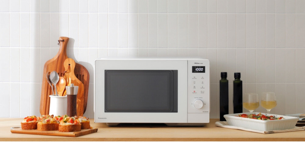 KUCHNIA MIKROFALOWA PANASONIC NN-GD34QWEPG Biała kuchenka mikrofalowa Panasonic ustawiona na drewnianym blacie przy jasnej kafelkowej ścianie, z deskami do krojenia i przekąskami po lewej stronie oraz oliwą, kieliszkami i naczyniem z potrawą po prawej