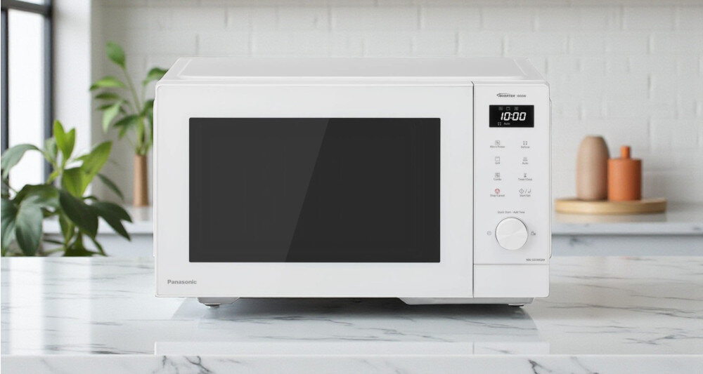 KUCHNIA MIKROFALOWA PANASONIC NN-GD34QWEPG  Biała kuchenka mikrofalowa Panasonic ustawiona na blacie z białego marmuru, na tle jasnej ceglanej ściany, z roślinami po lewej stronie i dekoracyjnymi naczyniami po prawej kompaktowy model GD34 funkcja grilla wydajne podgrzewanie grillowanie chrupiący kurczak zdrowe warzywa technologia inwerterowa Panasonic 14 automatycznych programów intuicyjne pokrętło łatwa obsługa szybkie gotowanie