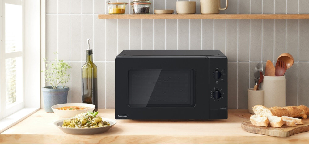 KUCHNIA MIKROFALOWA PANASONIC NN-SM22QBEPG Czarna kuchenka mikrofalowa Panasonic z pokrętłami mechanicznej regulacji stoi na drewnianym blacie jasnej kuchni, otoczona daniami, pieczywem i akcesoriami, podkreślając jej prostotę obsługi i estetyczne dopasowanie do wnętrza