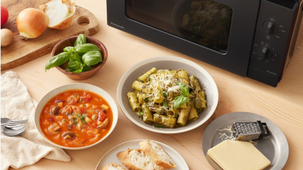 KUCHNIA MIKROFALOWA PANASONIC NN-SM22QBEPG Zbliżenie na blat z potrawami ustawionymi wokół czarnej kuchenki mikrofalowej Panasonic: talerz makaronu z pesto, miska z zupą warzywną, świeża bazylia, pieczywo oraz ser z tarką, co podkreśla wszechstronne zastosowanie urządzenia do podgrzewania i przygotowywania różnorodnych dań posiłki o każdej porze śniadanie obiad kolacja 5 poziomów mocy 200 W 800 W łatwe rozmrażanie szybkie podgrzewanie przygotowanie dań bez wysiłku