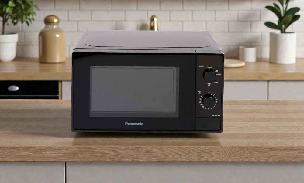 KUCHNIA MIKROFALOWA PANASONIC NN-SM22RBEVG Zdjęcie przedstawia czarną kuchenkę mikrofalową Panasonic ustawioną na białym blacie w jasnej kuchni. Wokół urządzenia umieszczone są składniki i potrawy, m.in. warzywa, przyprawy i gotowe danie. Po prawej stronie widoczne są rośliny doniczkowe na parapecie. Kompozycja podkreśla funkcjonalność sprzętu i jego estetyczne dopasowanie do małych wnętrz.