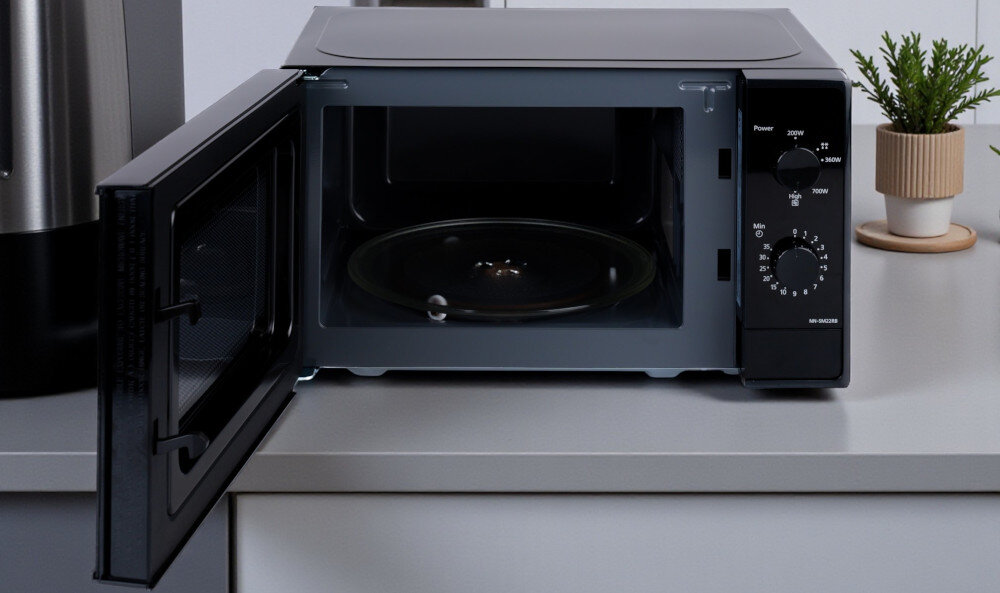 KUCHNIA MIKROFALOWA PANASONIC NN-SM22RBEVG Zdjęcie przedstawia czarną kuchenkę mikrofalową Panasonic z otwartymi drzwiczkami, ustawioną na jasnym blacie w kuchni. Wewnątrz widoczny jest szklany talerz obrotowy. Po prawej stronie znajduje się doniczka z zieloną rośliną, a po lewej fragment innego kuchennego sprzętu wnętrze emalia łatwoczyszcząca gładka powierzchnia łatwe czyszczenie talerz obrotowy równomierne podgrzewanie wygodne wykończenie urządzenie do podgrzewania potraw