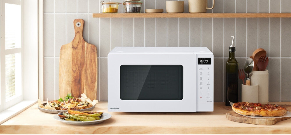KUCHNIA MIKROFALOWA PANASONIC NN-GT21QWEPG Biała mikrofalówka Panasonic z panelem dotykowym i wyświetlaczem LED, ustawiona na jasnym blacie kuchennym w otoczeniu deski do krojenia, oliwy i potraw, tworząca przytulną aranżację