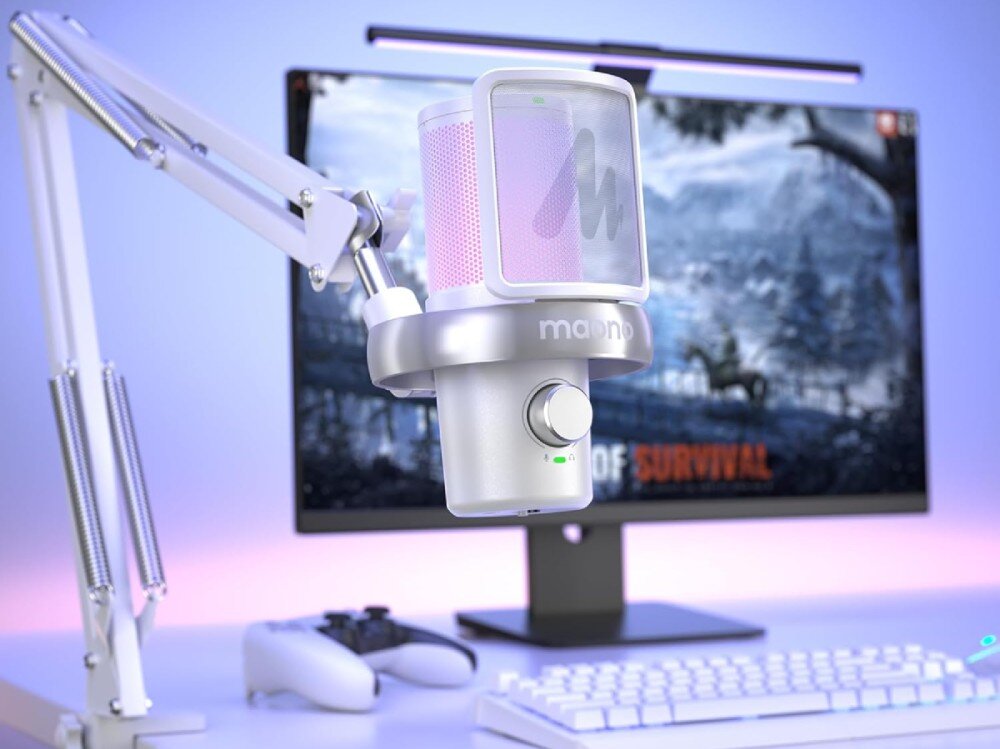 Na obrazie widoczny jest biały uchwyt do mikrofonu z logo \'maono\', zamontowany na biurku z monitorem, klawiaturą i kontrolerem do gier. W tle na ekranie monitora wyświetlana jest gra z napisem \'OF SURVIVAL\'.