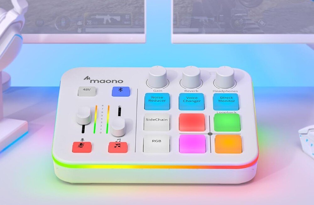 Kontroler do produkcji muzycznej i podcastów MAONO G1 Neo biała karta dźwiękowa z kolorowymi przyciskami i pokrętłami stoi na biurku przed dwoma monitorami wyświetlającymi grę komputerową, a spód urządzenia zdobi świecący pasek rgb