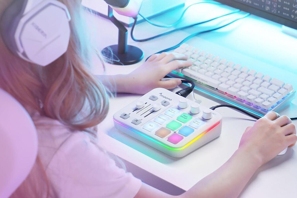 Kontroler do produkcji muzycznej i podcastów MAONO G1 Neo osoba w białych słuchawkach siedzi przy biurku i obsługuje jasny kontroler dźwięku z kolorowym podświetleniem, mając przed sobą białą klawiaturę oraz myszkę