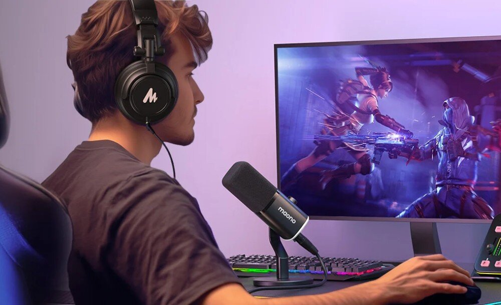 Mikrofon MAONO PD100X Mezczyzna w czarnych sluchawkach siedzi przed monitorem komputera, na ktorym wyswietlona jest dynamiczna scena z gry wideo. Na biurku obok klawiatury stoi czarny mikrofon na statywie, skierowany w strone twarzy uzytkownika