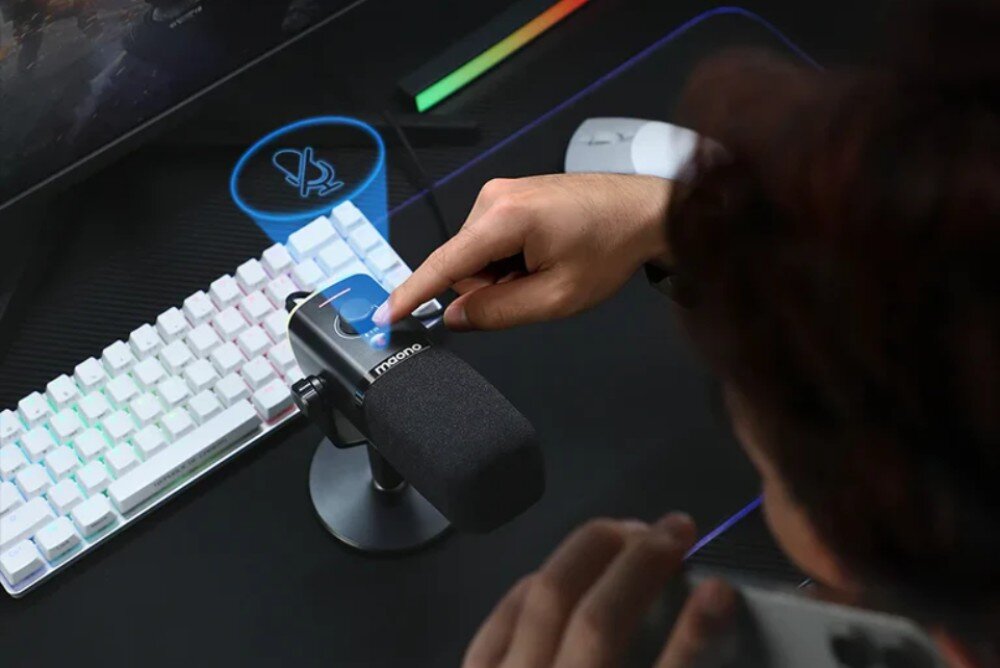 Mikrofon MAONO PD100X Widok z gory na czarny mikrofon stojacy na biurku obok bialej klawiatury. Palec wskazujacy dotyka gornej czesci obudowy, nad ktora unosi sie niebieska grafika przedstawiajaca przekreslony symbol mikrofonu