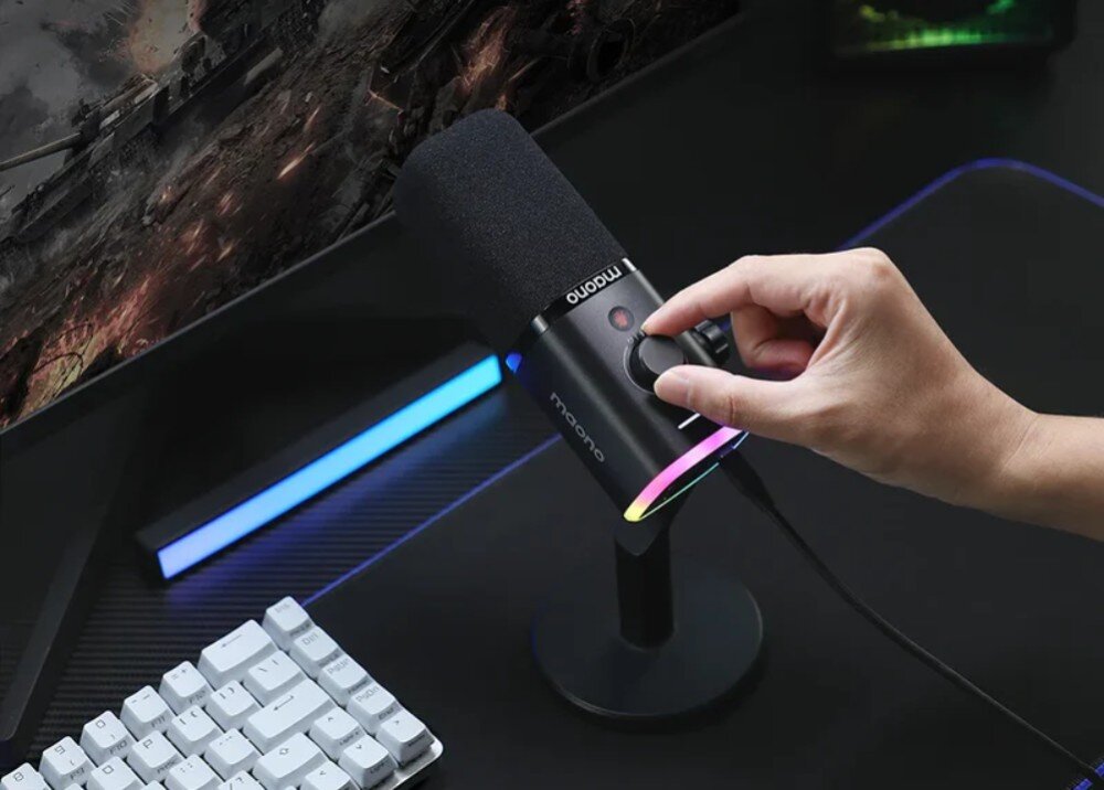 Mikrofon MAONO PD100X Mezczyzna reguluje czarne pokretlo umieszczone na obudowie mikrofonu. Urzadzenie stoi na statywie biurkowym, a w tle widoczny jest fragment monitora oraz podswietlana listwa dekoracyjna