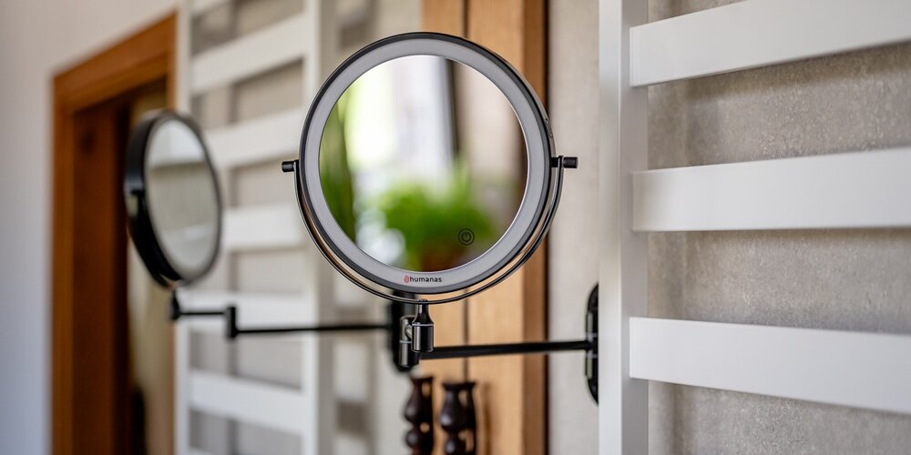 Okrągłe czarne lustro Humanas na regulowanym ramieniu, zamontowane na ścianie, z ikoną przycisku na tafli; w tle lustro ścienne