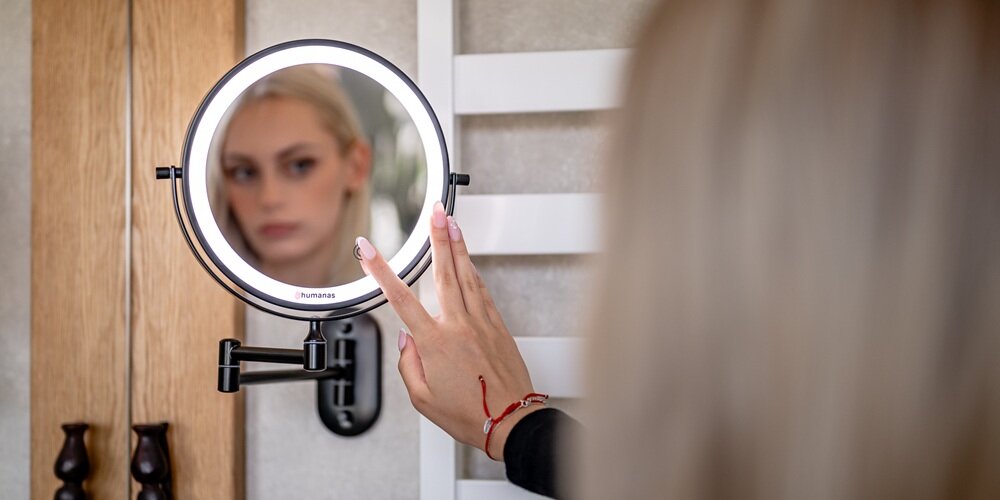 Okrągłe czarne lustro Humanas z podświetlanym pierścieniem na ramieniu ściennym, w odbiciu twarz; dłoń dotyka przycisku na tafli