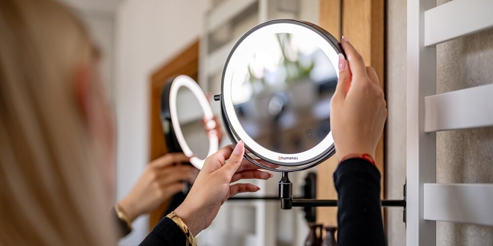Kobieta reguluje okrągłe lustro Humanas z podświetlanym pierścieniem na ściennym ramieniu; w tle odbicie drugiej strony lustra
