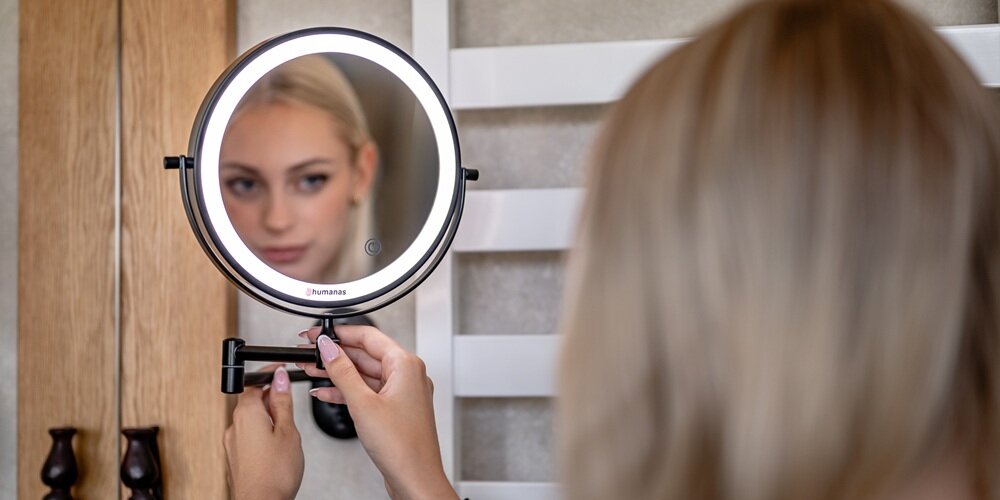 Okrągłe czarne lustro Humanas z podświetlanym pierścieniem na ramieniu ściennym, odbija twarz kobiety i jej dłonie