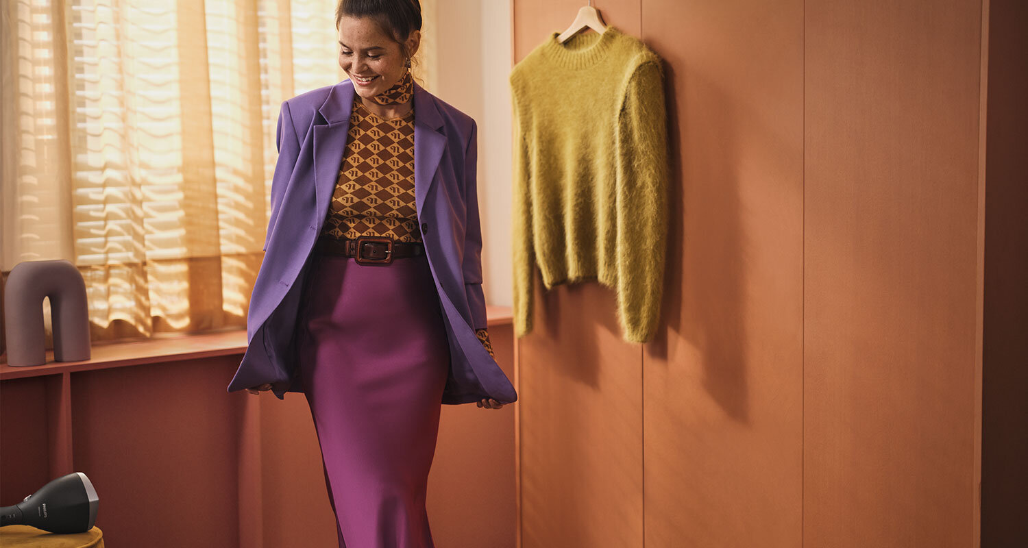 Kobieta w fioletowym żakiecie i spódnicy stoi w pomieszczeniu z drewnianymi panelami, obok wisi żółty sweter. Na półce znajduje się parownica do ubrań.