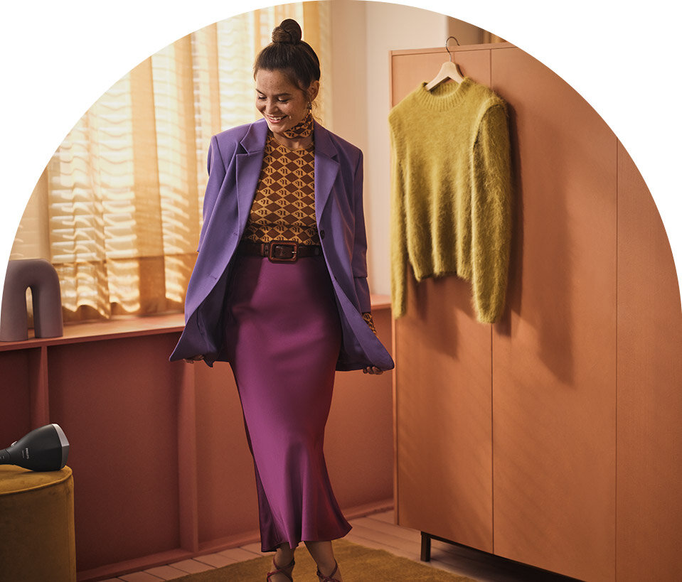 Osoba ubrana w fioletową marynarkę i spódnicę stoi w pomieszczeniu z drewnianą szafą i żółtym swetrem na wieszaku. Na podłodze widoczna jest parownica do ubrań.