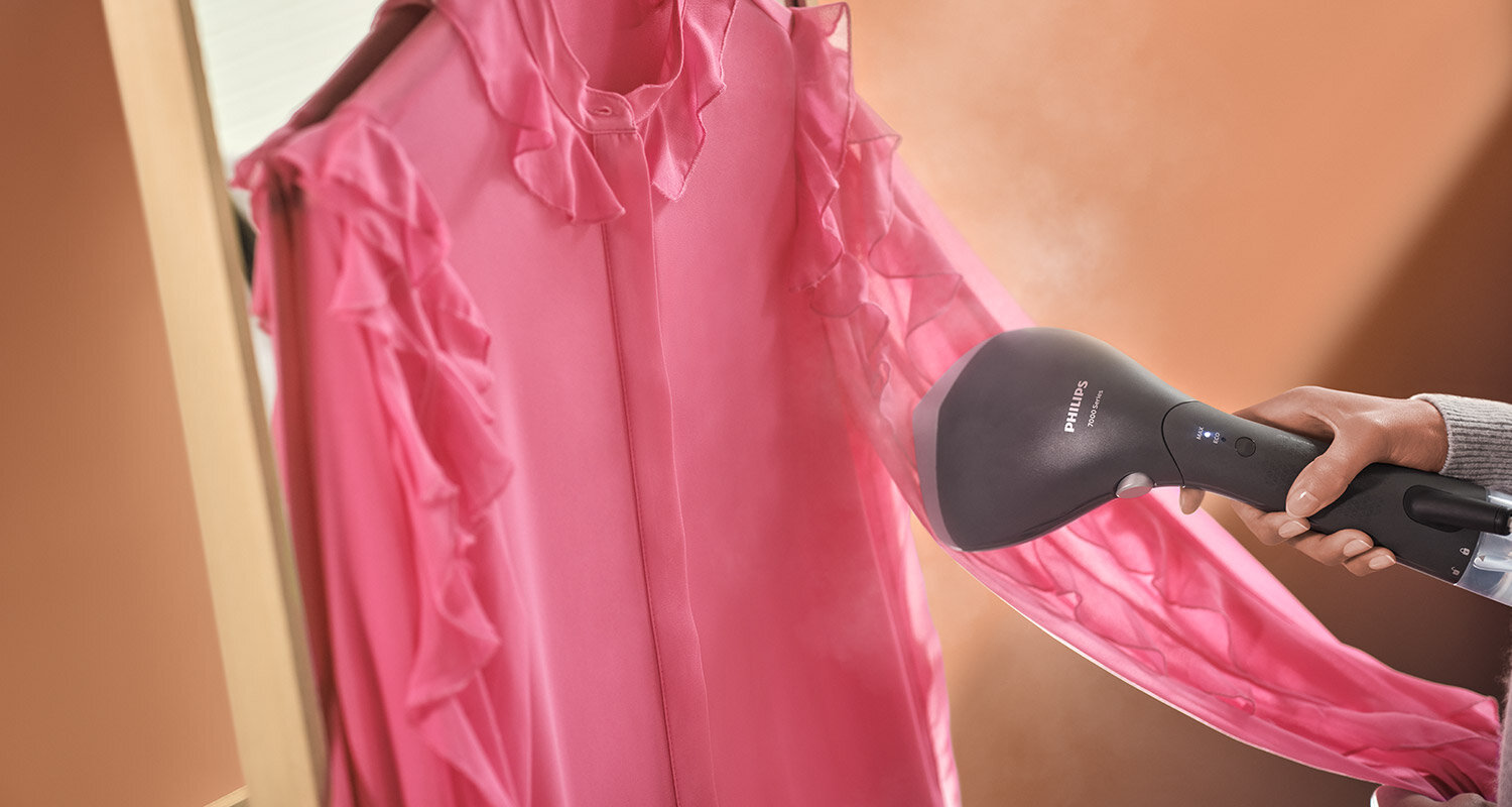 Osoba używa parownicy do ubrań na różowej koszuli z falbanami. Na urządzeniu widoczne jest logo \'PHILIPS\'.