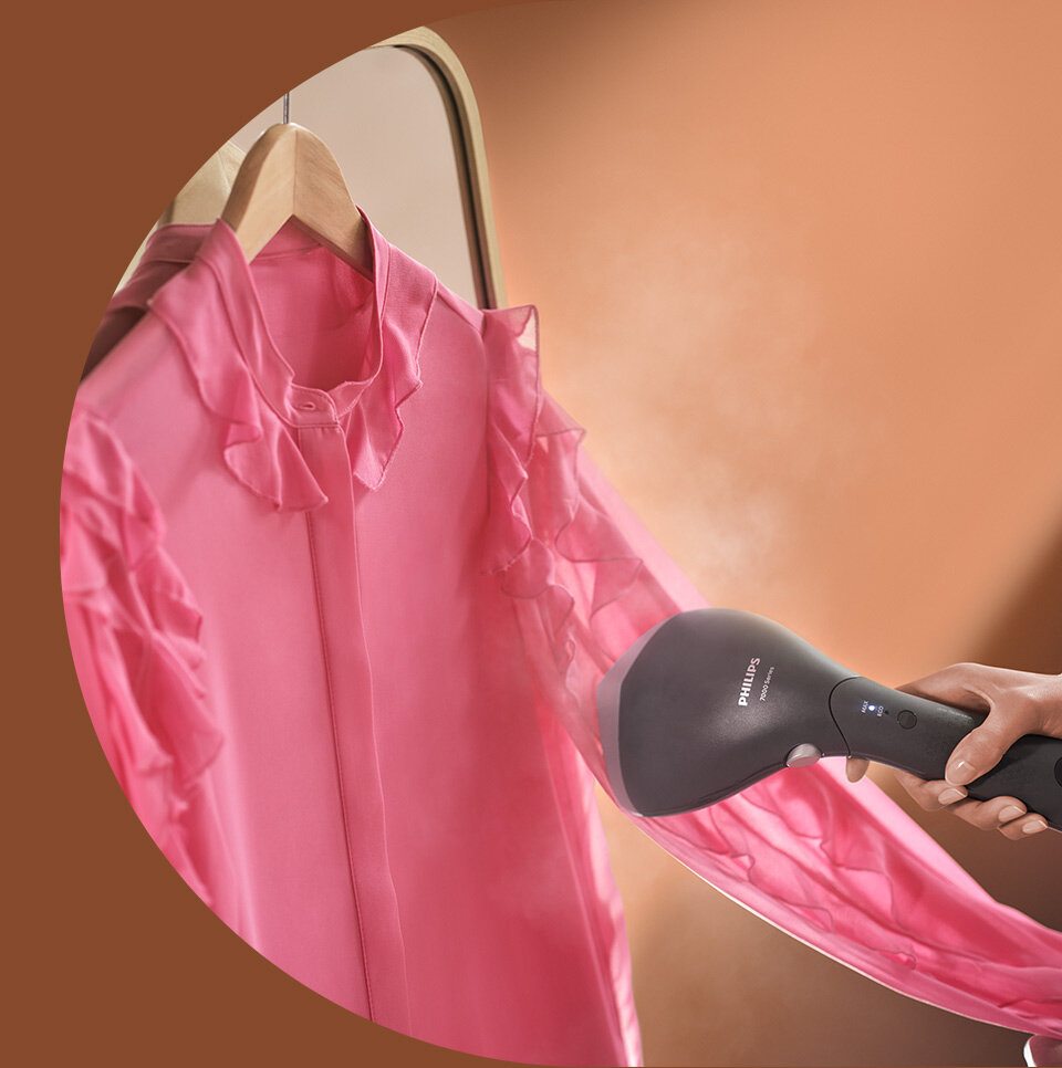 Osoba używa parownicy do ubrań na różowej koszuli wiszącej na wieszaku. Na parownicy widoczne jest logo \'Philips\'.