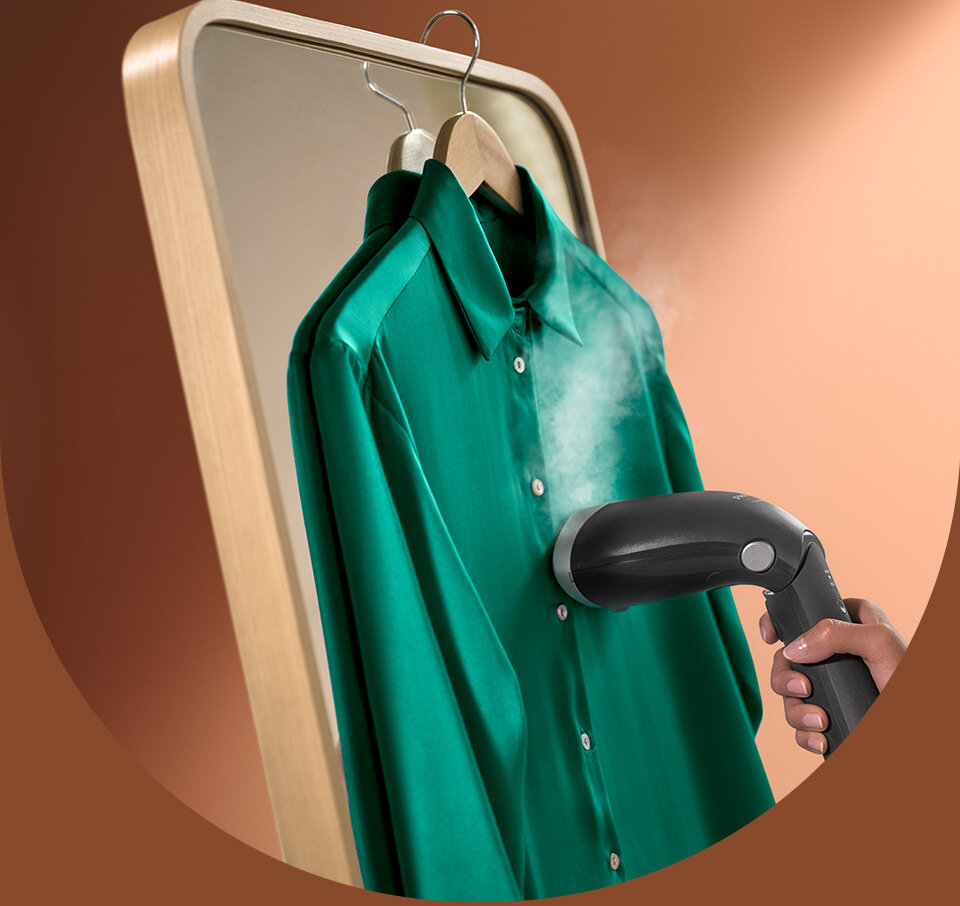 Osoba używa parownicy do ubrań na zielonej koszuli wiszącej na wieszaku. Z urządzenia wydobywa się para, a koszula jest umieszczona przed lustrem w drewnianej ramie.