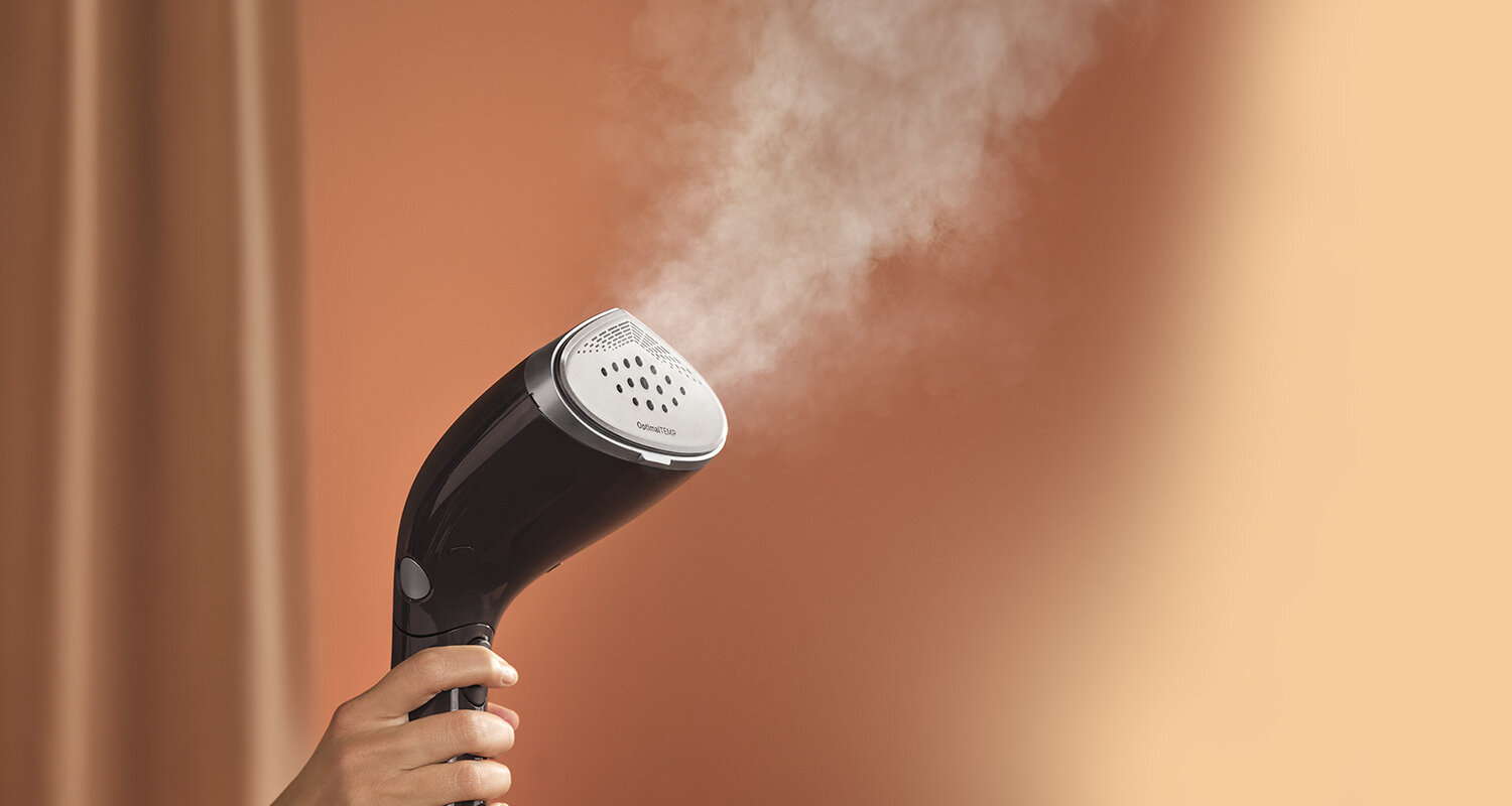 Osoba trzyma parownicę do ubrań, z której wydobywa się para. W tle widoczna jest gładka, brązowa ściana.