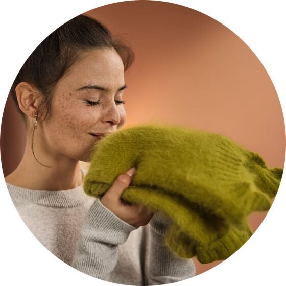 Osoba trzyma w rękach zielony, puszysty sweter i zbliża go do twarzy. Tło jest w ciepłym, brązowym odcieniu.