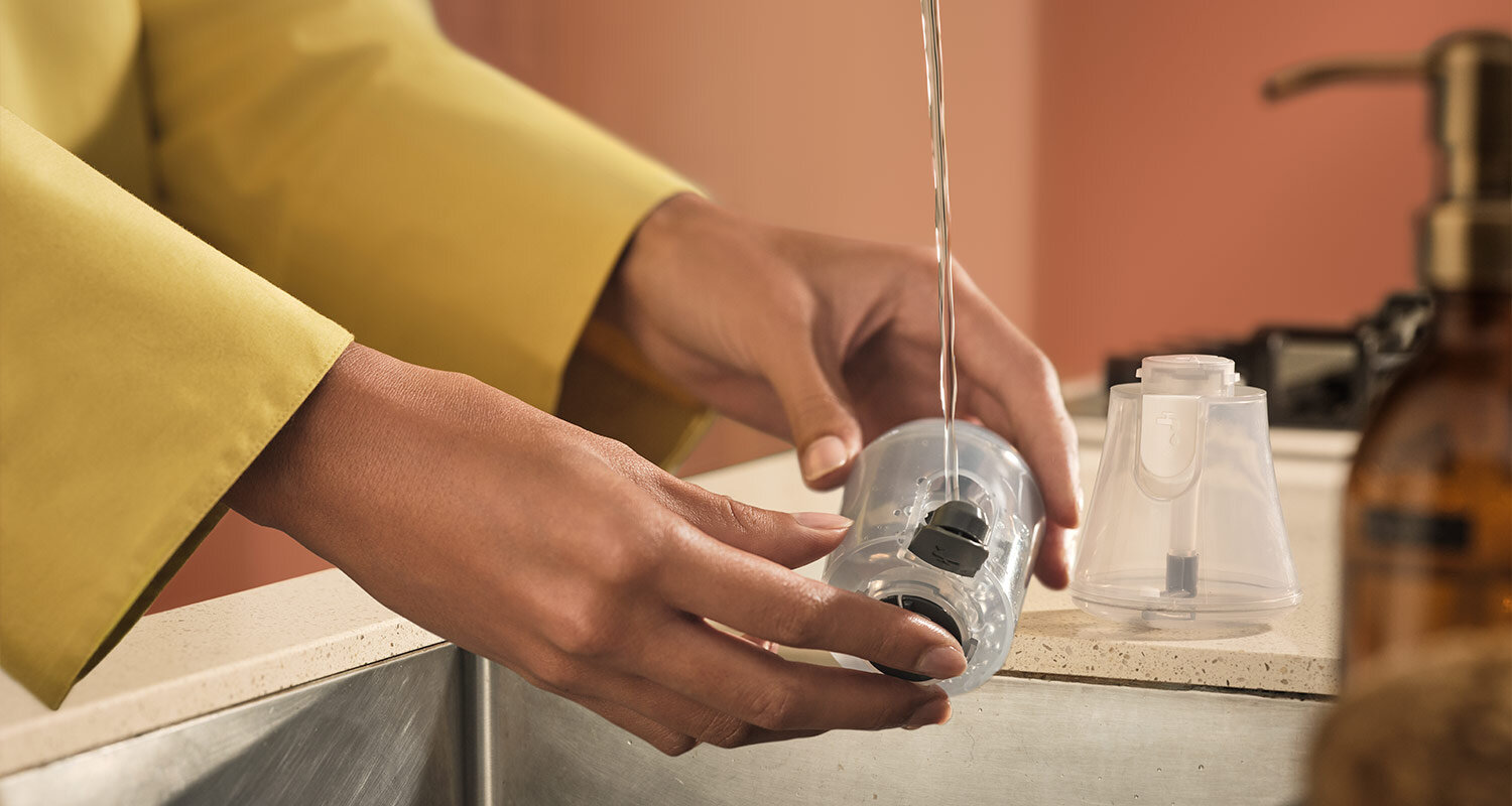 Osoba napełnia wodą przezroczysty pojemnik nad zlewem. Obok znajduje się drugi, podobny pojemnik oraz butelka z pompką.