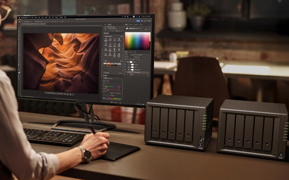 Serwer plików SYNOLOGY DS1525+ Osoba siedząca przy biurku korzysta z komputera z dużym monitorem, na którym otwarty jest program graficzny, obok stoją dwa czarne urządzenia sieciowe z kilkoma zatokami na dyski, opis