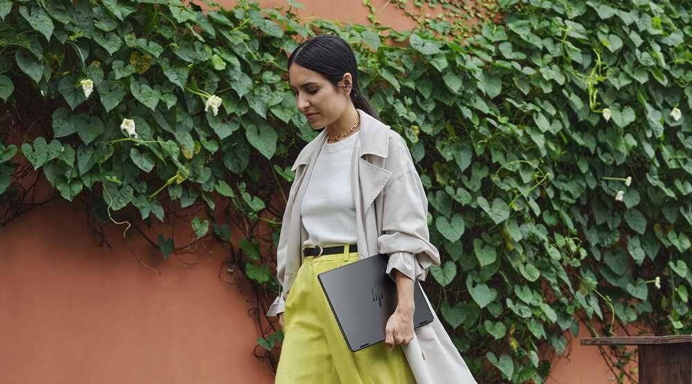 Kobieta w żółtych spodniach i białej koszuli idzie ulicą, trzymając laptop HP OmniBook Ultra Flip. Zrównoważony rozwój EPEAT Gold z Climate 