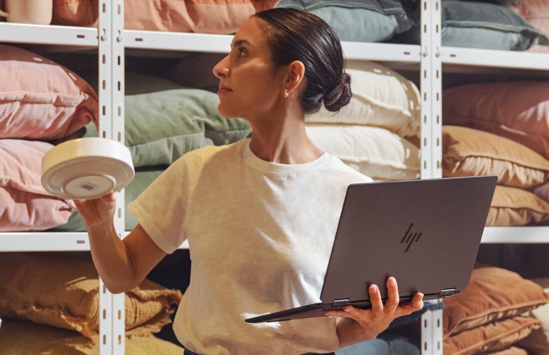 Kobieta trzymająca laptop HP OmniBook Ultra Flip przed półką z poduszkami.  Intel Core Ultra wspierany przez AI