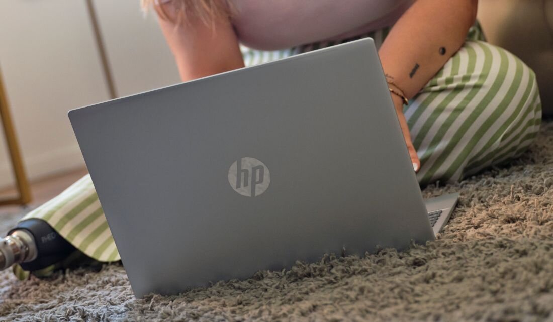 Laptop HP OmniBook z zamkniętą pokrywą leżący na dywanie obok kobiety w pasiaste spodnie - Moduł TPM