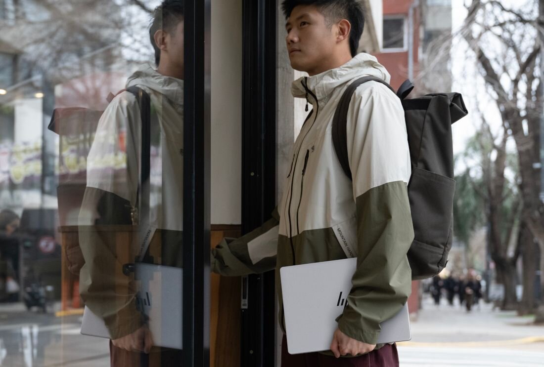 Mężczyzna w beżowej kurtce z plecakiem trzymający srebrny laptop HP OmniBook przy witrynie sklepowej. - Pamięć LPDDR5  