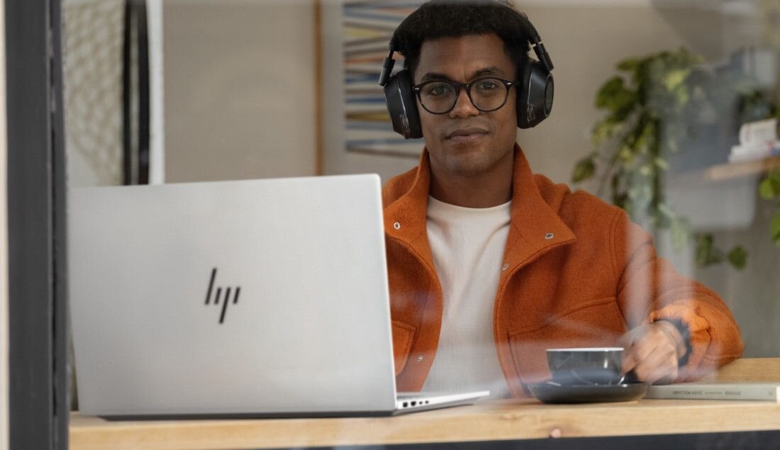 Mężczyzna w pomarańczowej kurtce i słuchawkach pracujący na laptopie HP OmniBook w kawiarni, podkreślający wysoką jakość dźwięku dzięki technologii DTS:X Ultra i komfort pracy zdalnej. - Głośniki z technologią DTS:X Ultra