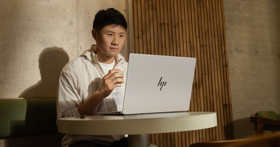 Mężczyzna w białej bluzie pracujący na srebrnym laptopie HP OmniBook przy okrągłym stole z drewnianą ścianą w tle. - Opis serii OmniBook 7