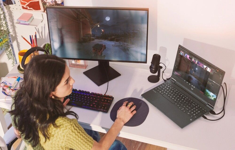 Kobieta w żółtym swetrze grająca na laptopie HP Victus 15 w domowym biurze z zewnętrznym monitorem, klawiaturą mechaniczną i mikrofonem. 