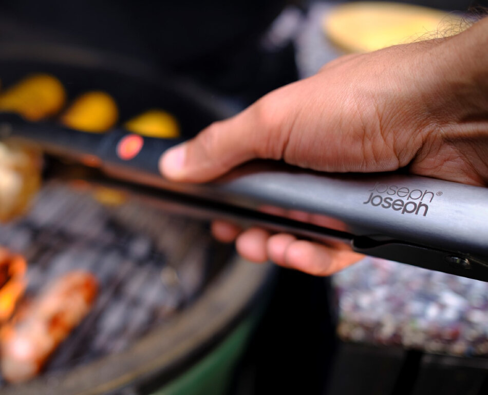 Osoba trzyma metalowe szczypce grillowe z widocznym logo \'Joseph Joseph\'. W tle widać grill z jedzeniem.