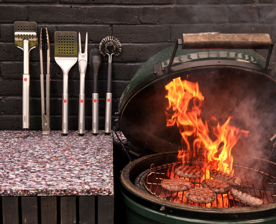 Na obrazie widoczne są narzędzia grillowe ustawione na blacie oraz otwarty grill z płomieniami i grillowanym mięsem. Narzędzia mają metalowe uchwyty z czerwonymi elementami.